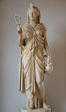 Marble staue of a woman holding a rattle in one hand and a pitcher in the other.