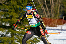 Photographie d'un biathlète en combinaison noire, portant des lunettes et des bâtons relevés vers le haut.