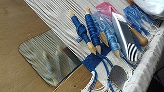 Tapestry tools on a loom: mirror, bones (wrapped with yarn), scraper (with short teeth), heavy comb (double-ended, to batten the weft), and awl (tip hidden).