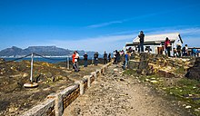 A straight stone pathway through a rocky area, elevated in places from the walkway, with a low retaining wall and chain fence on the left. Beyond is an area of ocean; at the far background on the left is a large flat rocky mountain with a peaked one at its left. Closer to the camera is a small building on the right; people are milling around it and the paths, some taking pictures