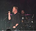 A male singer, wearing a black shirt and holding a microphone, is singing. A drummer is performing behind him.