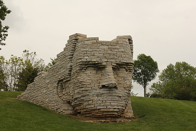 The 12-foot sculpture on a grassy hill with trees in the background is a portrait of Leatherlips, made from stacks of flat, rectangular-shaped limestone rocks. The stacked stones form the shape of Leatherlip's face, then extend in a straight line behind the head until the hill on which it is built. This part is an enclosure in which people can stand and take in the same view as Leatherlips: the river and an amphitheater.
