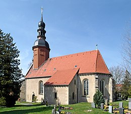 Béžová budova kostela z boku, červená střecha, hnědá věž, vlavo strom, v popředí náhrobky, v pozadí modrá obloha