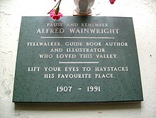 black stone plaque reading: Pause and remember Alfred Wainwright: Fellwalker, guide book author and illustrator who loved this valley. Lift you eyes to Haystacks his favourite place. 1907–1991