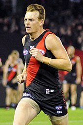 A male athlete with light hair wearing a sleeveless jersey and shorts runs on the grass surface of the playing arena.