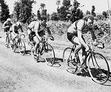 Plusieurs cyclistes, vus de côté, en file indienne, se déplaçant de gauche à droite.