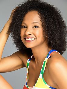 Indhira Serrano wearing a patterned sleeveless top, seated with elbow on one knee, head turned sideways, grinning at camera