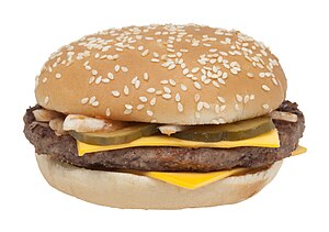 A brown meat patty between slices of yellow American cheese, topped with sliced pickles and lettuce, sandwiched in a sesame seed bun