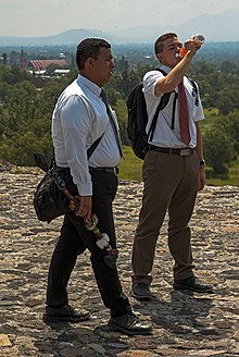 Two young men, the one on the left darker-skinned than the one on the right, who is drinking some orange liquid from a plastic bottle, standing on a stone surface with mountains in the background. They are both carrying backpacks and wearing white dress shirts, ties and slacks. The one on the right is wearing a short-sleeved shirt and tan slacks