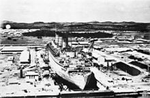 An ocean liner, viewed from aft and above, sits in a dry dock.