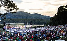 View of an amphitheater in front of a river with rolling hills in the background, with West Point cadets in front of the stage and performance attendees in the foreground