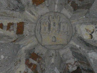 Photo en couleur d'une clef de voûte en pierre entre des briques formant nervures. La clef est sculptée d'un cercle contenant un château à trois tours.