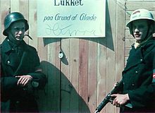 Closed due to happiness. Two Danish resistance fighters are guarding a shop while the owner is celebrating the liberation of Denmark on 5 May 1945. The man on the left is wearing a captured German Stahlhelm, the one on the right is holding a Sten gun. Closed due to happiness.jpg