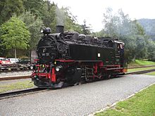 Kouřící černě zbarvená parní lokomotiva řady 99.73 stojící na kolejích u vydlážděného nástupiště. V pozadí jsou lesy.