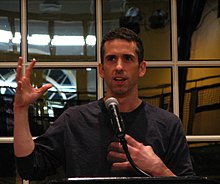 A middle-aged white man with dark hair, wearing a dark top, talking in front of a microphone