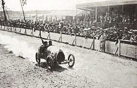 Photo d'une voiture en course avec son pilote et son mécanicien embarqué passant à vive allure devant la foule.