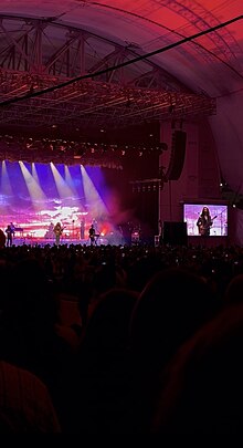 The image shows Hozier's stage from a distance during the Unreal Unearth tour at Leader Bank Pavilion in Boston, MA. Hozier is on stage performing Work Song on the left of the image. The backdrop shows a pink and purple sunset with various spotlights shining down on Hozier. A screen zoomed in on Hozier is to the right of the stage. Red and pink lights project across the venue matching the color scheme of the backdrop.