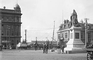 A black and white photograph of a town square with several people standing near a statue of Queen Victoria in the middle.