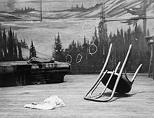 An overturned chair in front of a mural, on which several chalk circles have been drawn around bullet-holes