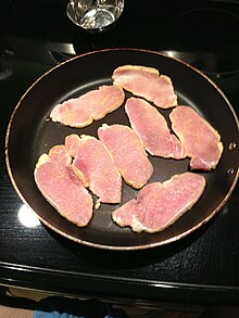 black frying pan on black stovetop, with eight slices of peameal bacon