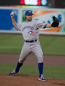 A man wearing a gray baseball uniform with blue socks, "Toronto" written across the chest in blue letters, and a blue cap prepares to throw a baseball from the pitcher's mound