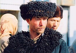 A photograph of a Uyghur man standing. He is wearing a hat and sporting a goatee.