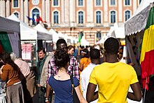 Photographie couleur montrant des personnes marchant entre des stands couverts de chapiteaux blancs, avec en toile de fond la façade du Capitole de Toulouse.