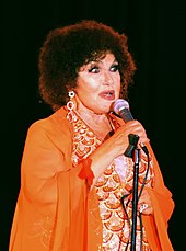 A woman wearing an orange dress and earrings, holding a microphone.