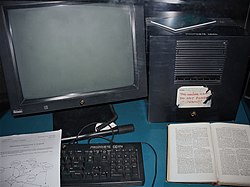 Black equipment on a teal blue desk. At left a monitor and at right a cube, both with small NeXT logos and in front a keyboard that says "Propriete CERN". Resting on the keyboard is a copy of "Information Management: A Proposal," and to its right is a book, probably "Enquire Within upon Everything". A partly peeled off label on the cube says, "This machine is a server. DO NOT POWER IT DOWN!!"