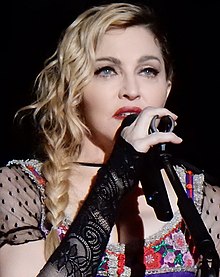 A closeup photo of Madonna with shoulder-length wavy blonde hair, wearing a colorful, low-cut blouse, holding a microphone to her mouth with her right hand.