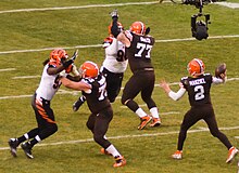 Photographie d'une action de football américain lors de laquelle un joueur est en train de lancer un ballon, protégé par plusieurs joueurs de ligne.