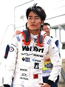 Photographie d'un jeune homme japonais, brun, vu de face, en plan américain, habillé dans une combinaison de pilote blanche.