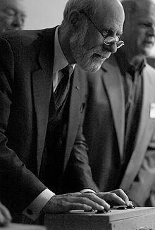 Homme portant des lunettes et un costume, légèrement penché en avant et regardant au loin, avec les mains posées sur une manette de jeu et appuyant sur des boutons.