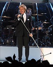 Мужчина на сцене в костюме держит микрофон и подставку под углом. Позади него мужчина играет на барабанной установке.