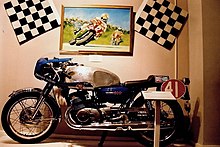 Race prepared road bike with race number 41, a large shiny aluminium fuel tank and streamlined top half fairing with dropped handlebars in front of a wall mounted large painting of Mike Hailwood in 1978 on a TT race prepared Ducati leading Phil Read on a Honda flanked by twin chequered flags