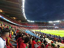 Seitliche Farbfotografie einer vollen Stadiontribüne mit Zuschauern in rot-grünen Kleidern. Auf dem Dach steht „UEFA Euro 2016“. Rechts sitzen mehrere Sicherheitskräfte mit Blick auf die Tribüne und weiter rechts sitzen Fotografen aneinandergereiht mit Blick auf das Spielfeld. Im Hintergrund sind das Stadion mit roten und schwarzen Punkten und die Spieler auf dem Rasen zu sehen.
