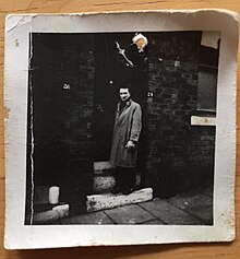 A young Roger Frety standing on the front steps of his row house fraternity in a black and white 1950s photograph in a long brown overcoat.