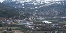 Luftfoto einer in bergiger Umgebung liegenden Ortschaft. Die Berge im Hintergrund sind teils mit Schnee bedeckt.