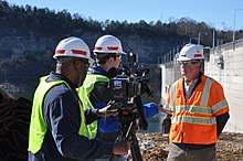Two men in hard hats, one holding a camera, interview another man in a hard hat at a construction site. The camera has a Fox 17 logo sticker on it.