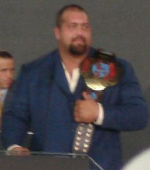 un homme portant une veste bleu avec une ceinture de champion de catch sur l'épaule gauche.
