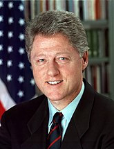 A presidential photograph of a Caucasian man wearing a suit with a black tie. An American flag and bookshelf are in the background.