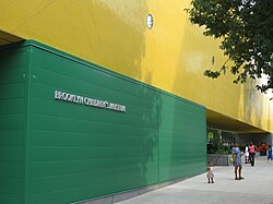 A green exterior wall stands one story high, and atop it, the wall is yellow. On the green portion are the words "BROOKLYN CHILDREN'S MUSEUM" in silver font. Some adults and children stand on the sidewalk in front of the building.