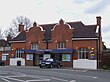 A brown-bricked building with a black roof and a blue sign that reads "CHIGWELL STATION" in white letters all under a white sky