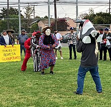A group of dancers in a grassy field, with audience nearby, and a sign reading "Danza del Torito de Romita Guanajuato" in back; one dancer is wearing a bull mask with horns; one dancer is wearing a dress and a mask with a woman's round face; two more dancers are behind her in a black-and-white striped suit and a red costume.
