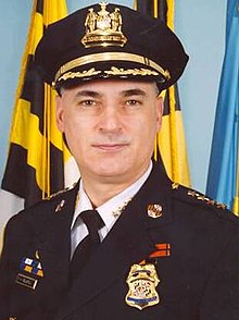 Bealefeld, in front of a Maryland, Baltimore, and unidentified blue flag, wearing a black suit jacket with a Baltimore Police Department badge, a nametag which reads "F. H. BEALEFELD III", and various other badges, over a white uniform with a black tie, along with a peaked police cap.