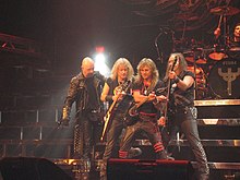 The band Judas Priest are onstage at a concert. From left to right are the singer, two electric guitarists, the bass player, and the drummer, who is seated behind a drumkit. The singer is wearing a black trenchcoat with metal studs.