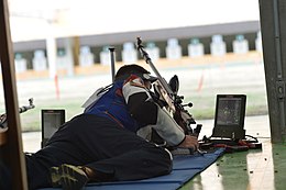 Photograph of a man lying down to shoot a rifle, with full shooting jacket and trousers, a shooting mat and electronic target display.