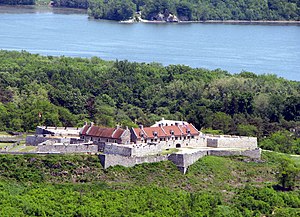 The star-shaped fort is visible in the center of the photograph, with its inner buildings roofed in red. The fort is surrounded by forest, and a body of water (a portion of Lake Champlain) is visible behind the fort.