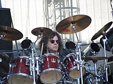 Zamračený Jiří Pátek s rozpuštěnými kudrnatými vlasy a černými brýlemi sedí za červenou bicí soupravou