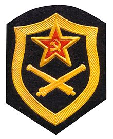 A black patch with a yellow outline of a shield embroidered on it. Inside the shield, at the top, there is a red star with a yellow hammer and sicle inside. Below that, there are two yellow rockets crossing over each other.